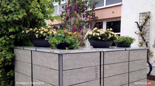 Ein modernes Garten-Q Flachdach-Gerät mit blühenden Pflanzen oben, umgeben von Büschen und Gebäude im Hintergrund.