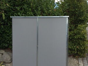 . The image shows a gray metal outdoor storage cabinet placed on a concrete surface. It's situated in front of some greenery, likely plants or shrubs, with a partly cloudy sky in the background.