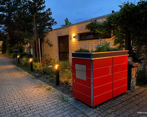 Ein rotes Paketlager mit Pflanzen; beleuchteter Weg und Gebäude im Hintergrund, umgeben von Bäumen und Büschen.