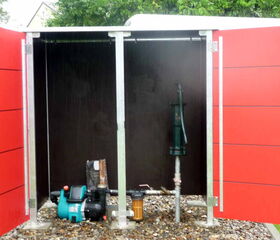 . The image shows an open enclosure with red doors. Inside, there is a green pump and some plumbing equipment on a gravel surface. It appears to be used for water management or irrigation purposes.