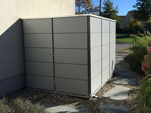 . Ein graues Metall-Gartenhaus an der Hauswand, umgeben von Kies und einem Gartenweg.