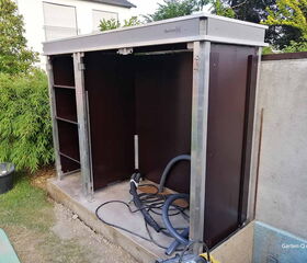 Ein Garten-Unterstand mit Metallrahmen und Regalen, Kabeln am Boden, neben einem Haus und Bambus.