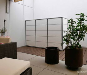 . The image depicts a minimalist outdoor patio area. Key elements include:

1. **Storage Unit**: A sleek, white storage cabinet against the wall.
2. **Seating**: Dark wicker outdoor furniture with light-colored cushions.
3. **Plants**: Two potted plants, one on a table and another larger plant on the floor.
4. **Flooring**: Combination of tiled and wooden flooring.
5. **Wall and Covering**: Plain white walls with a retractable awning above.
6. **Decor**: A small table with a floral cover and a plant pot.

The space is designed to be functional and aesthetically pleasing, suitable for a relaxing outdoor environment.