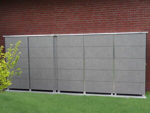 . Ein grauer Gartenschrank steht vor einer roten Ziegelwand. Daneben ist grüne Vegetation sichtbar.