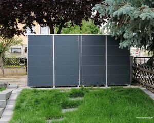 . The image shows a garden storage unit or shed with a modern design, featuring a sleek, dark gray exterior. It's placed in a garden area with grass and paving stones, surrounded by trees and a wooden fence.