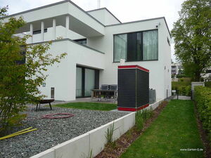 . This image shows a modern white house with a flat roof. There is a garden area with gravel and grass, along with some shrubs or small trees. A patio area with chairs and a table is also visible. In the foreground, there's a tall, narrow structure with a red top, possibly a storage unit or utility box.
