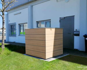 Ein modernes Holz-Verkleidungselement steht neben einer Gebäudewand mit Tür und Fenstern auf einer Wiese.
