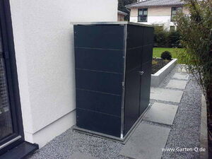 . Metallschrank neben einem Haus auf einer Kiesfläche, hinter einem Weg und Gartenbeet.