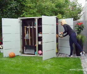 . The image shows a person organizing or retrieving items from an outdoor storage cabinet or shed. The cabinet is open and contains various garden tools and sports equipment like rakes, shovels, brooms, and balls. There is also a basketball on the grass. The surroundings include lush greenery and some potted plants with red flowers.