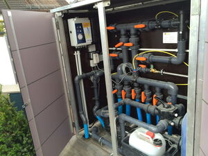 . The image shows the interior of a cabinet containing a complex arrangement of plumbing pipes and valves. The pipes are primarily made of PVC and include several orange-handled valves for controlling the flow of water or another fluid. There are also some electrical components and wiring present, which may be part of a control system for the plumbing setup. A white container with a red cap is seen at the bottom, likely holding a liquid used in the system. This setup is typically found in systems like water filtration or pool management.