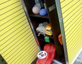 . The image shows the interior of a yellow cabinet or locker with several shelves. Here's what's visible in the image:

1. **Top Shelf**: Various containers and objects.
2. **Middle Shelves**: A bag, a toy truck, and a few other items.
3. **Bottom Shelf**: A small red toy car with a white steering wheel, partially outside the cabinet.
4. **Floor**: The surface outside the cabinet appears to be ground or soil.

The cabinet has a metal frame and doors.