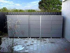. The image shows a backyard with a large, gray storage unit or shed. It's set against a fence with trees visible above the fence line. The ground is paved with tiles, and there’s some tall grass or plants in the foreground on the left side. There's a circular cover on the ground in front of the storage unit.