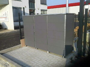 . The image shows a large, rectangular gray structure, possibly a storage unit or utility box, situated outdoors on a paved surface. It appears to have a metallic finish and is secured on four raised supports. The surrounding area includes industrial buildings and a fenced perimeter.
