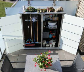 Ein geöffneter Schrank auf einem Balkon mit Gartenutensilien und Dekorationen oben drauf. Vorne ein Tisch mit Blume.