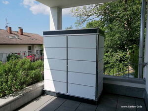 . The image shows a modern outdoor storage cabinet or shed situated on a patio or balcony. It's sleek and modular in design with a white finish and dark trim. There is greenery and a residential building in the background.