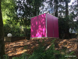. This image shows a modern, pink-colored garden shed placed in a wooded outdoor setting. The area is surrounded by trees and greenery, creating a natural backdrop. There's a white spherical light fixture on the left side, adding to the garden's aesthetic.
