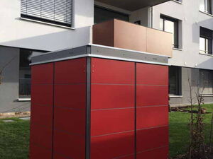 . The image shows a red shed-like structure placed in front of a modern apartment building. The shed has a metallic frame and is covered in red panels. The apartment building is contemporary, with multiple floors and balconies, and there is grass and paving surrounding the shed.