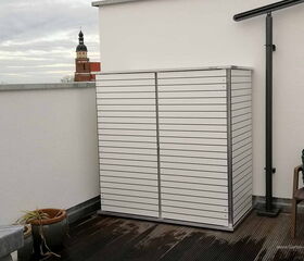 Ein kleiner, weißer Gartenschrank auf einer Terrasse, mit einer Pflanze in einem Topf und einem Kirchturm im Hintergrund.