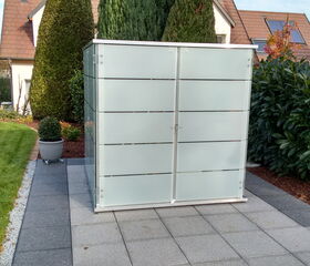 . The image shows a modern, frosted glass enclosure situated on a paved area in a garden. There are large hedges and shrubs around, along with a well-maintained lawn. The setting appears to be a residential backyard.