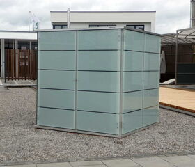 . This image shows a glass and metal structure with a modern design. It is located on a gravel and paved area, possibly an outdoor space adjacent to buildings. The structure has opaque glass panels and is surrounded by other architectural features, including a wooden deck and additional buildings.