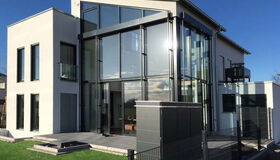. The image shows a modern building with a sleek, minimalist design. The structure features large glass windows and a combination of concrete and metal elements, providing a contemporary aesthetic. The building appears to be a house situated in a yard with a small lawn and paved area. There's also a metal enclosure or box in the foreground near a fence.