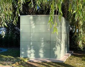 Ein modernes, grünes Gartenhaus unter hängenden Ästen, umgeben von Bäumen und Gras.