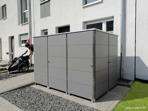 . Eine Person zieht einen Kinderfahrradanhänger aus einer grauen Fahrradgarage nahe eines Gebäudes.