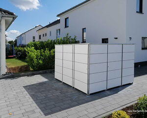 . The image shows a modern, white cube-shaped structure placed on a paved driveway. It's likely a storage unit or shed. The area around it includes a pathway with rectangular pavers, and there are residential houses with white walls in the background. Additionally, there are shrubs and a small lawn nearby, all under a clear blue sky.
