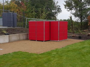 . The image shows three red metal structures with what appear to be doors, possibly lockers or storage units, arranged in an outdoor setting. They are positioned on a gravel area alongside a grassy lawn, with some trees and a fence in the background.