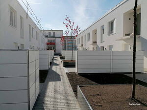 . Eine moderne Wohnanlage mit weißen Gebäuden, Zäunen und Bäumen entlang eines gepflasterten Wegs.