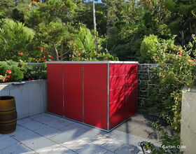 Ein roter Gartenschrank steht auf einer Terrasse, umgeben von Grünpflanzen und einer Steinmauer.