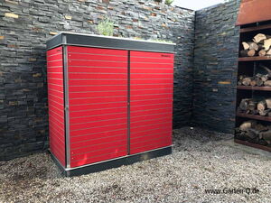 . The image features a red storage structure known as "Garten Q." It appears to be placed outdoors against a wall made of stacked stones. To the right, there's a wood rack containing cut logs. The ground is covered with gravel. The structure seems to be designed for garden or outdoor use.