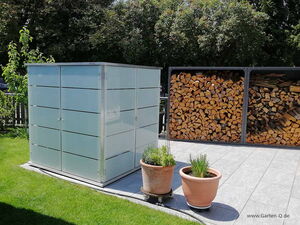 . The image shows a garden scene with a modern storage shed made of frosted glass or similar material on the left. To the right, there is a stack of firewood in a metal frame. In the foreground, there are two potted plants on a tiled patio. The surrounding area is lush with green grass and trees.