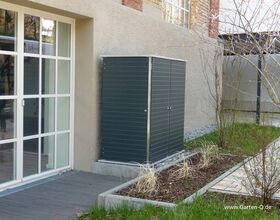 The image shows a dark-colored outdoor storage cabinet placed against the exterior wall of a building. The cabinet is beside a glass door leading outside. In front of the cabinet, there’s a small garden area with some plants and surrounding greenery. The ground is partly covered with wooden decking and there’s a pathway leading away from the area.