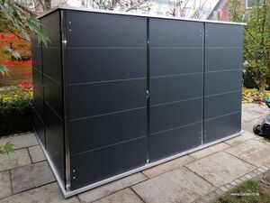 . Ein moderner Schuppen im Garten mit grauen Paneelen und umgeben von Pflanzen und Steinplatten.