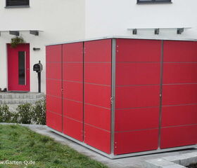 . The image shows a modern outdoor storage unit or shed with a sleek design featuring red panels and a metal frame. It is placed in a yard area, next to a house with a matching red door. The scene includes a small lawn, some paved walking areas, and a mailbox near the entrance of the house.