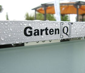 . The image shows a close-up of a metallic surface with water droplets on it. There is text on the surface that reads "Garten[Q]". In the background, there appears to be an outdoor structure with an orange canopy. The focus is mainly on the text and the water droplets.