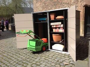 . The image shows an outdoor storage shed with its doors open. Inside the shed, there's a variety of gardening equipment and supplies, including:

1. A green lawn mower placed outside the shed.
2. Pots and planters on the shelves.
3. Some folded cushions or fabric items.
4. A bag of gardening soil or fertilizer.
5. A small hand shovel on one of the shelves.

The shed is located on a cobblestone surface and there are a few people in the background.