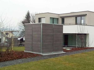 . The image shows a modern house with a cube-shaped structure in front of it. The cube appears to be covered with a wood-like material and has a metallic frame. The house has a flat roof, large windows, and is surrounded by a garden with sparse trees and grass. The setting appears to be residential, with overcast weather conditions.