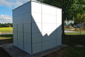 The image shows a cubic structure with a tiled exterior, located outdoors. It is situated on a paved area with grass and trees nearby, and a road can be seen in the background. The structure casts a distinct shadow, with some sunlight reflecting off one side.