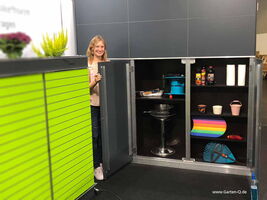 The image shows a storage cabinet with open doors. Inside, there are various items like a toolbox, cleaning supplies, and paper towels. A person is standing next to the cabinet with some plants on top. The cabinet exterior has bright green slats.