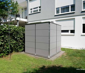 . The image features a modern, cube-shaped garden storage shed with a sleek gray exterior. It is situated on a small concrete platform in a yard with grass and near a hedge. In the background, there is a building with several windows and a balcony. There’s also a metal pole or rod next to the shed, likely part of a trellis or clothesline.