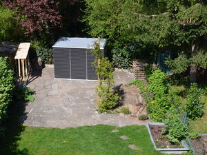 . The image shows a garden scene with a paved patio area and various plants and trees. There is a small structure that appears to be a shed or storage unit. Surrounding the patio are lush green plants, a vegetable garden, and a tree providing some shade. The area is well-maintained, with grass and decorative landscaping elements.