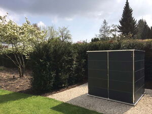 . The image shows a garden scene featuring a small, dark-colored shed or storage unit on a gravel area. There are green bushes and various trees in the background, including one with white blossoms. The sky appears to be partly cloudy.