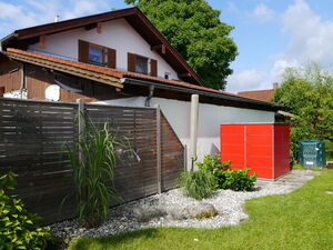 . The image shows a backyard scene featuring a house with a sloped roof and brown shutters. In the foreground, there's a wooden fence and a garden area with gravel, grass, and various plants. A red structure, possibly a shed or storage unit, is visible near the fence. There's also a green recycling or compost bin in the grass to the right.