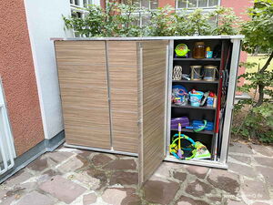 . The image shows an outdoor storage cabinet with shelves filled with various items. Here's a description of its contents:

1. Top Shelf: 
   - Watering cans
   - A green container
   - A brown jug

2. Second Shelf:
   - Lanterns
   - Other decorative items

3. Third Shelf:
   - Buckets with toys

4. Bottom Shelf:
   - Sports equipment (e.g., rackets)
   - Folded fabric or sheet

5. Tools and equipment are stored on the side.

The cabinet has a wooden appearance and is situated against a building wall.