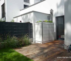 . Terrasse mit Holzboden, weißem Schuppen, schwarzem Zaun, Pflanzen und schlauchartigem Gartenwerkzeug.