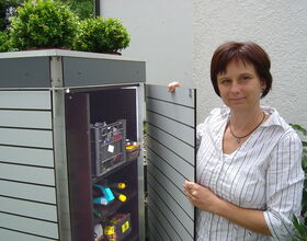 . The image shows a person standing next to an outdoor cabinet or storage unit with the door open. On top of the cabinet, there are small green plants in pots. Inside the cabinet, there are various tools and items stored on shelves, including a plastic crate and a handheld tool.