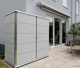 The image shows a modern outdoor storage unit located on a patio. It is placed near a table with chairs and close to a glass sliding door. The storage unit is sleek and contemporary in design, fitting the aesthetic of the outdoor space. The surrounding area includes potted plants and a well-maintained garden.