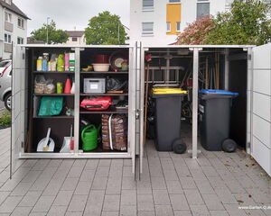 In the image, there is an outdoor storage unit with two compartments. The left compartment has shelves storing various items like cleaning supplies, gardening tools, and materials such as a watering can, bags of soil or fertilizer, and household items like a dustpan and brush. The right compartment contains large wheeled garbage bins, likely for waste management. The unit is located in a residential area with visible buildings and greenery in the background.